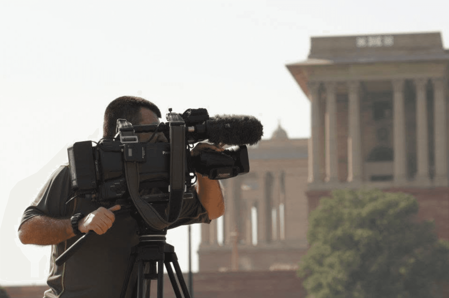 A camera operator filming outdoors with a large broadcast camera in front of an architectural backdrop